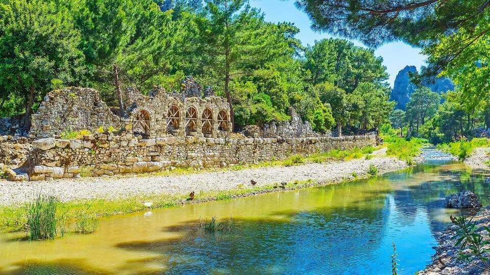 A river with a stone wall and arched windows, surrounded by lush green trees and a rocky riverbank.