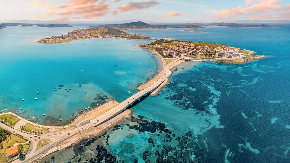 Une vue aérienne d’une ville côtière, avec un pont imposant reliant deux portions de terre, des eaux d’un bleu limpide et une île visible au loin.