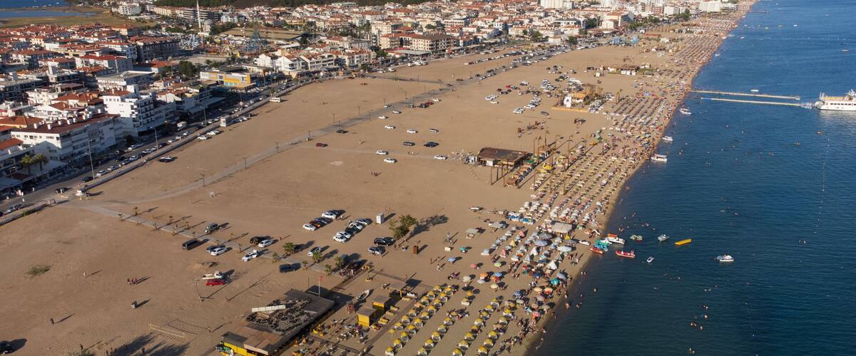 A coastal cityscape with a wide beach, parked cars, and a clear blue sea.