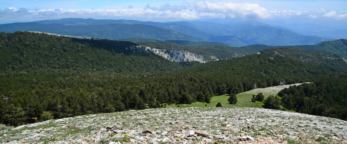A mountainous landscape with dense forests, rocky terrain, and distant mountain ranges under a clear sky.