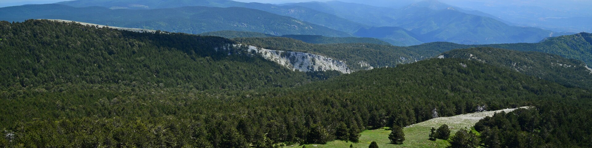 A mountainous landscape with dense forests, rocky terrain, and distant mountain ranges under a clear sky.