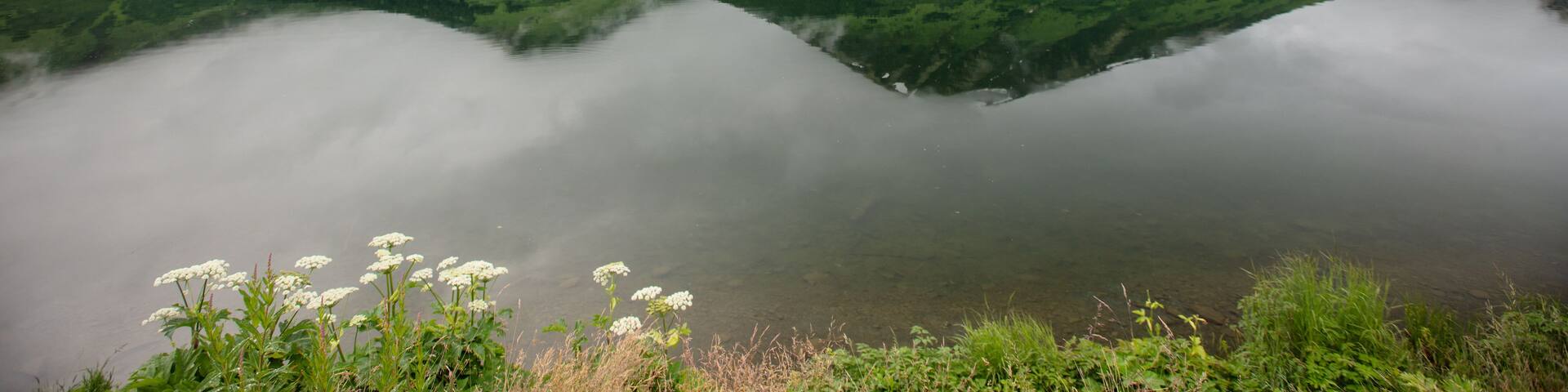 A calm lake reflecting a mountain range with lush greenery and a cloudy sky.
