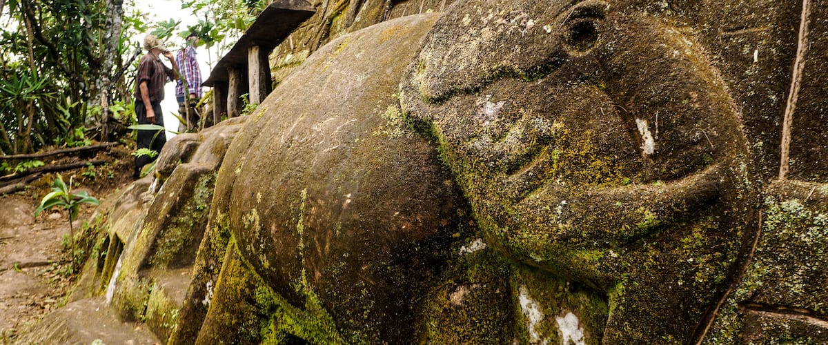 A large, moss-covered elephant statue with a carved face, set against a rocky cliff.