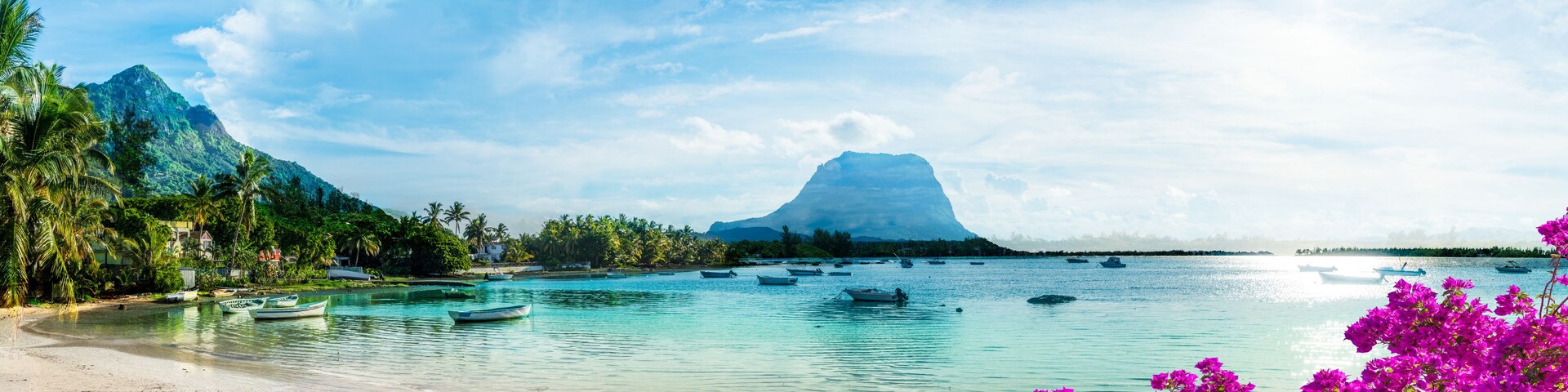 Una playa tropical con barcos, agua turquesa cristalina, palmeras y una montaña al fondo.