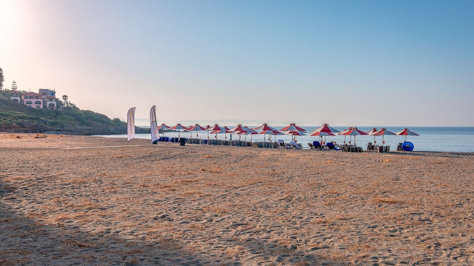 A beach with red and white umbrellas, a row of beach chairs, and a building in the background.