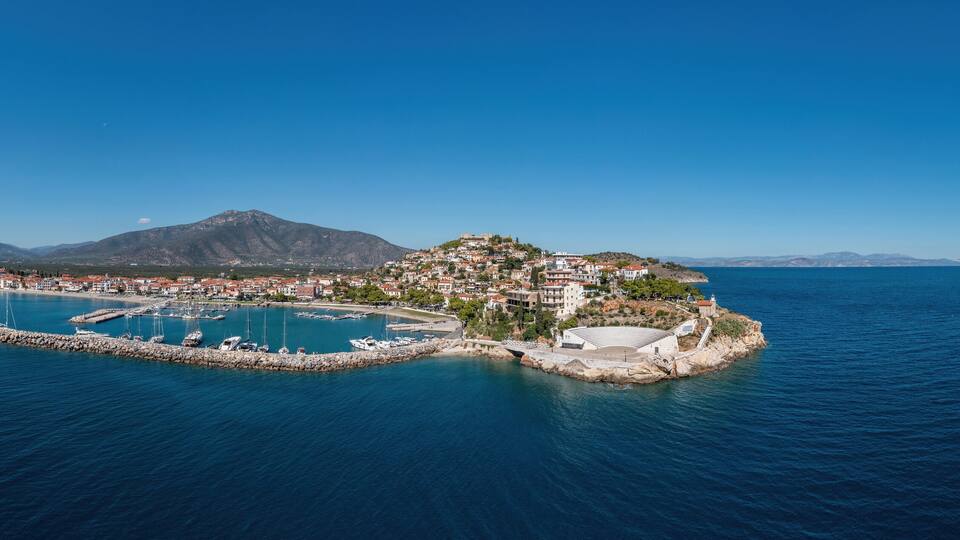 A coastal town with a marina, a small island with buildings, and mountains in the background.