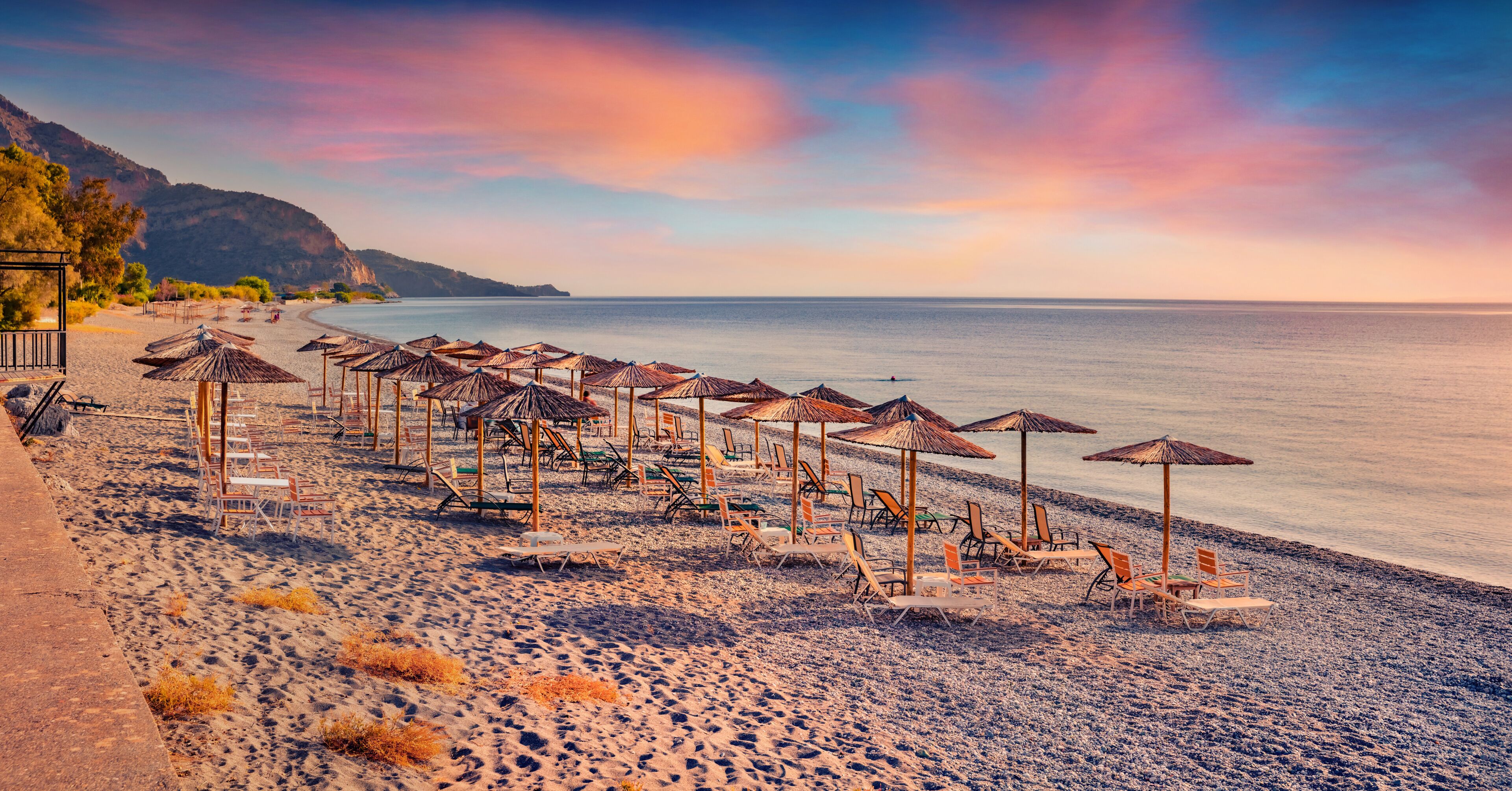 Une plage bordée de rangées de chaises longues et de parasols, une mer calme et un paysage montagneux en arrière-plan.