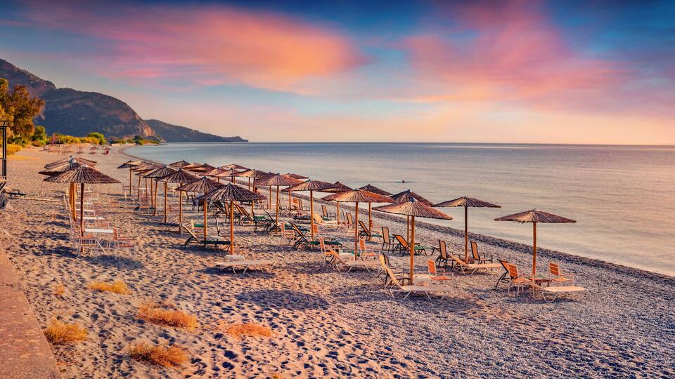 A beach with rows of sun loungers and umbrellas, a calm sea, and a mountainous backdrop.