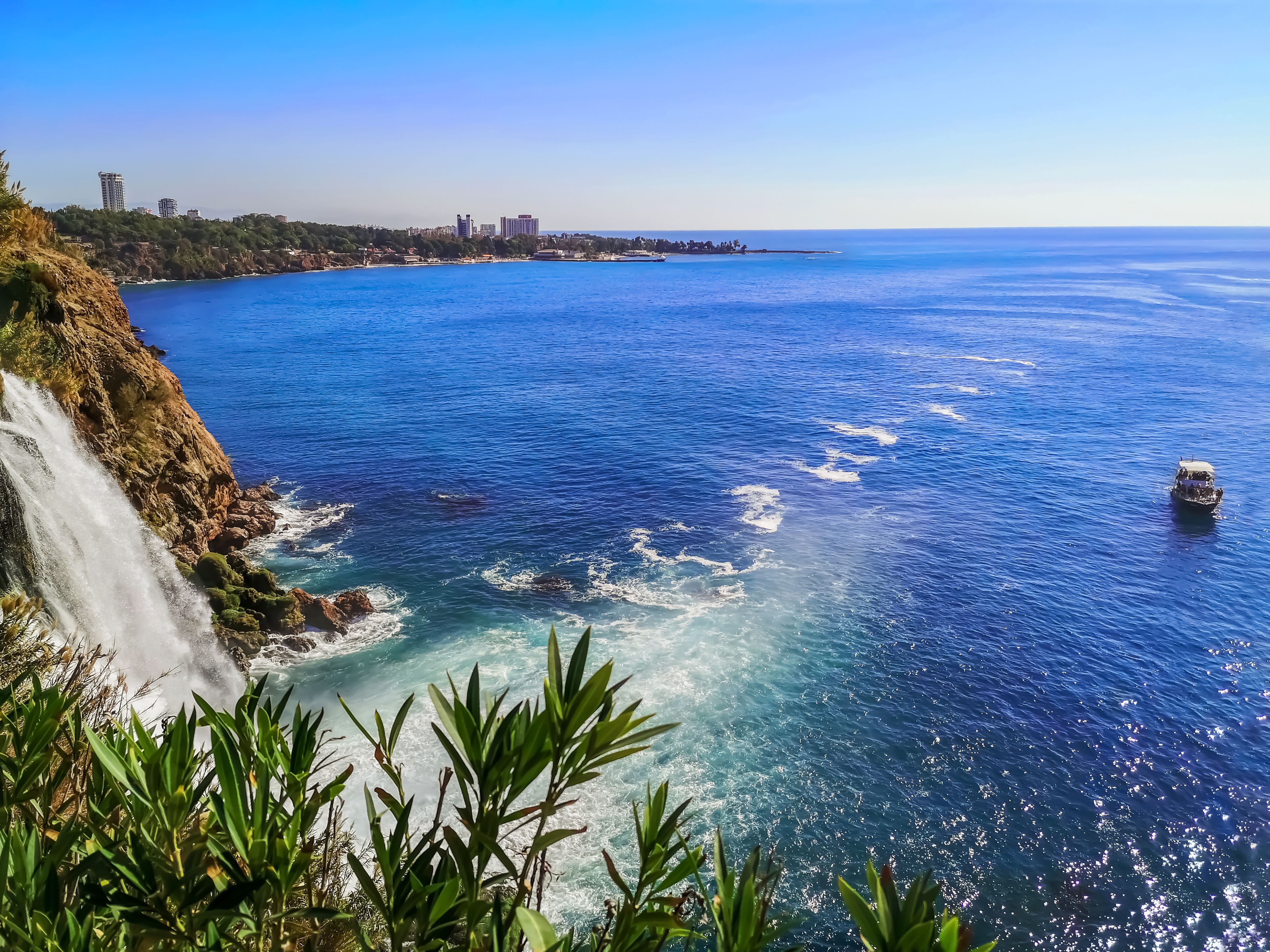 A coastal landscape with a clear blue sea, a rocky cliff with a waterfall, and a distant city skyline.