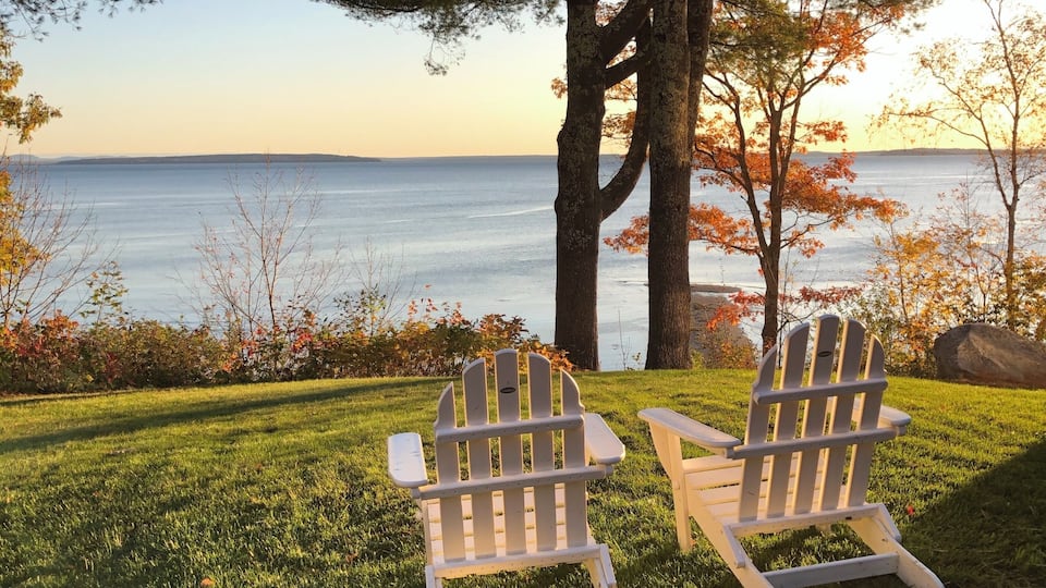 Two Adirondack chairs facing a lake with autumn foliage and a clear sky.