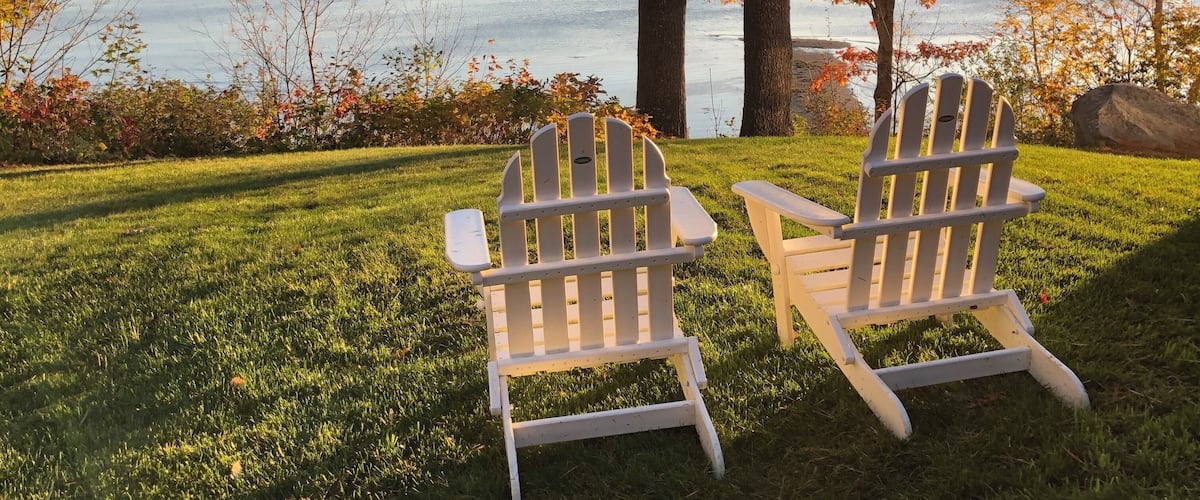 Two Adirondack chairs facing a lake with autumn foliage and a clear sky.