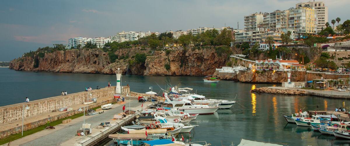 A marina with boats docked, a rocky coastline, and buildings in the background.