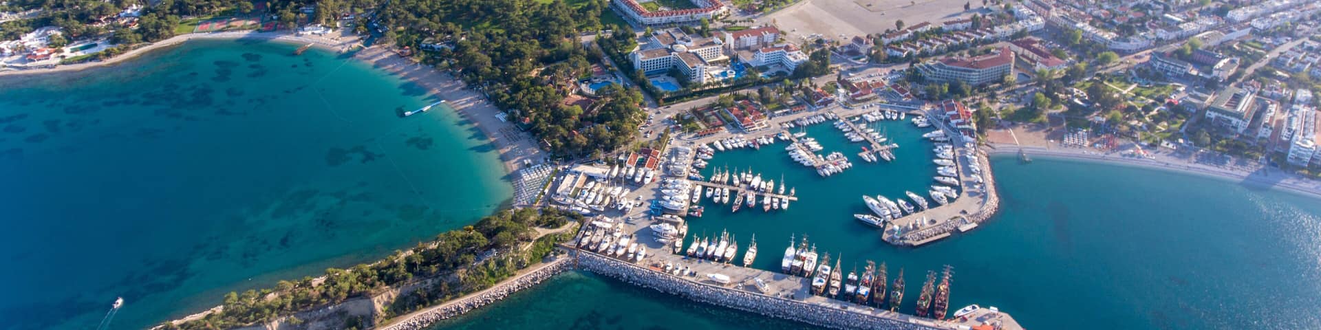 An aerial view of a coastal town with a marina, clear turquoise waters, and surrounding hills.