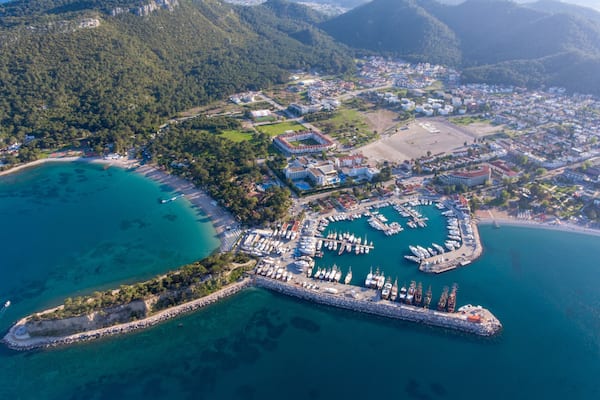 An aerial view of a coastal town with a marina, clear turquoise waters, and surrounding hills.