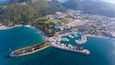 An aerial view of a coastal town with a marina, clear turquoise waters, and surrounding hills.