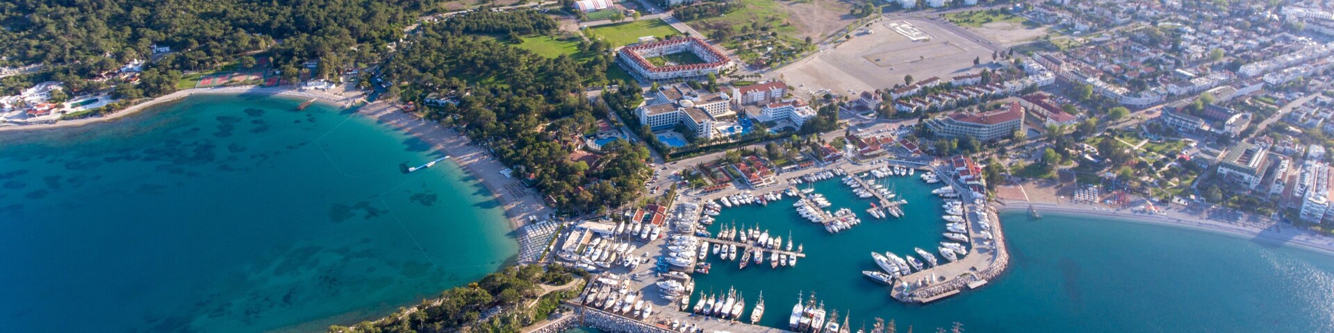 Vista aérea de un pueblo costero con un puerto deportivo, aguas turquesas cristalinas y colinas circundantes.