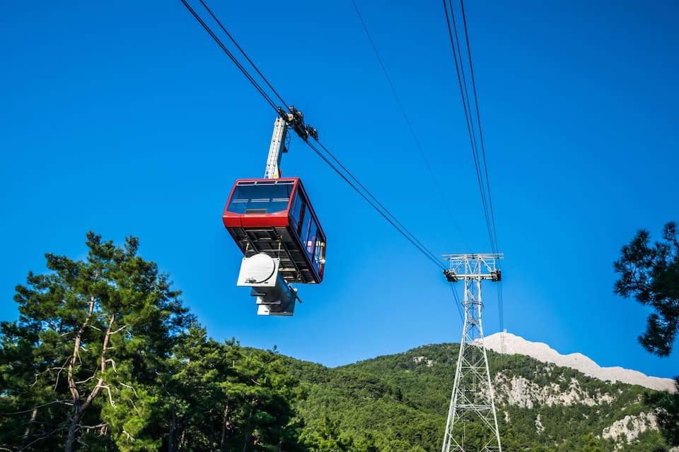 Une télécabine rouge suspendue entre deux tours, un ciel d’un bleu limpide et un paysage montagneux parsemé d’arbres.