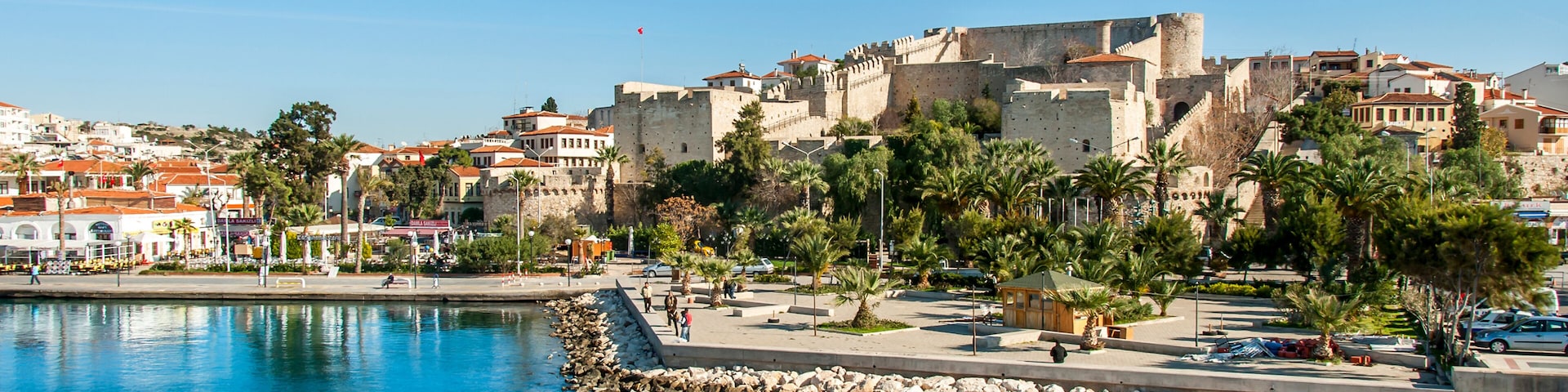 Un pueblo costero con un fuerte histórico, aguas azules y tranquilas, y un paseo marítimo bordeado de palmeras.