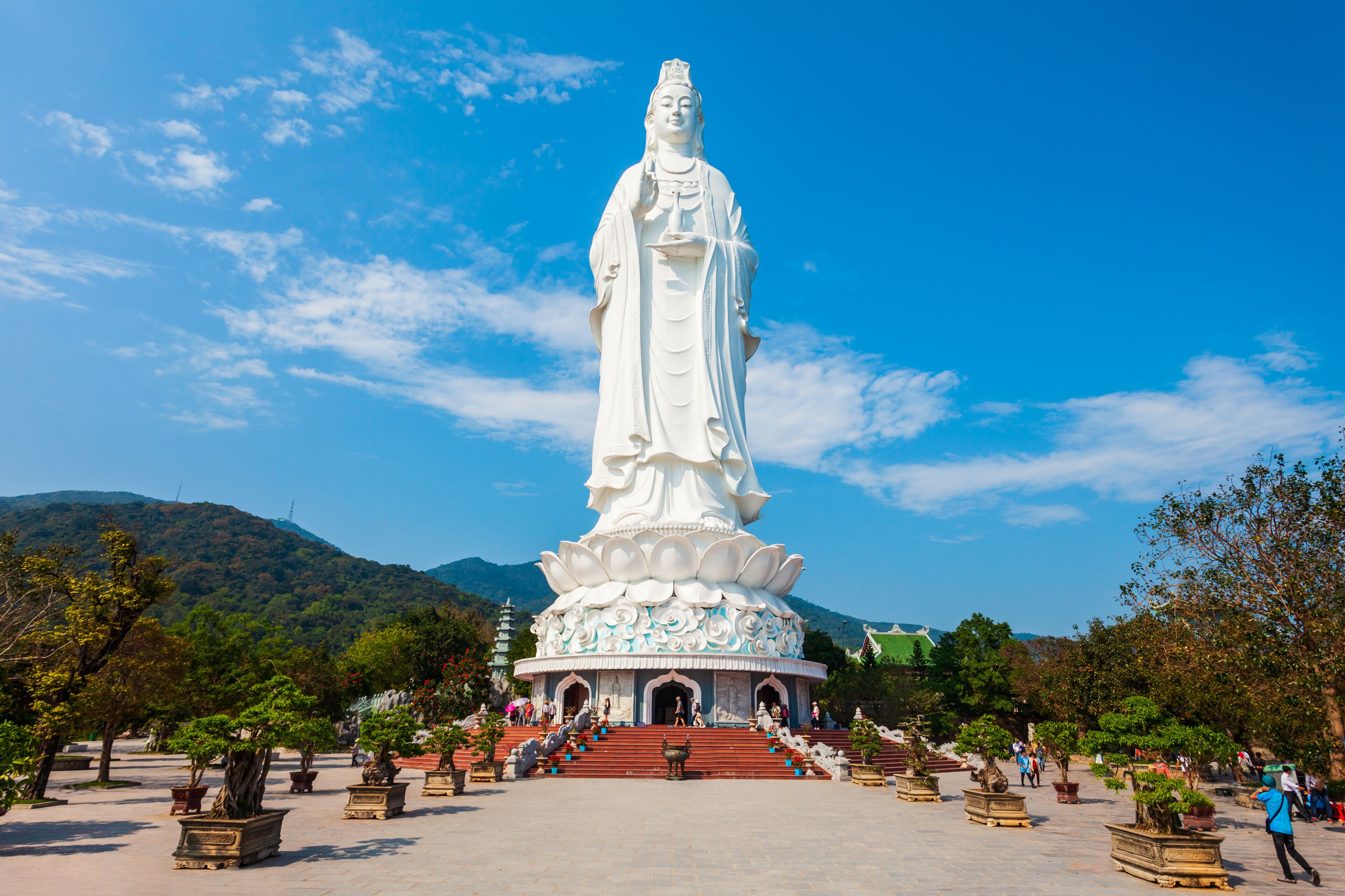 A large white statue with a lotus base, surrounded by potted plants and stairs leading up to it.
