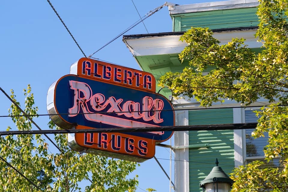 A neon sign for Alberta Rexall Drugs against a backdrop of a green building and trees.