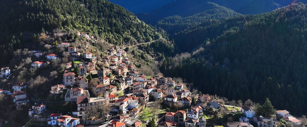 A mountainous village with houses on a hillside, surrounded by dense forests and a prominent snow-capped peak in the background.