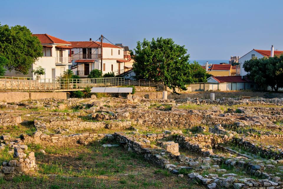 Ancient ruins with stone walls and scattered stones, a bridge, and houses with red roofs.