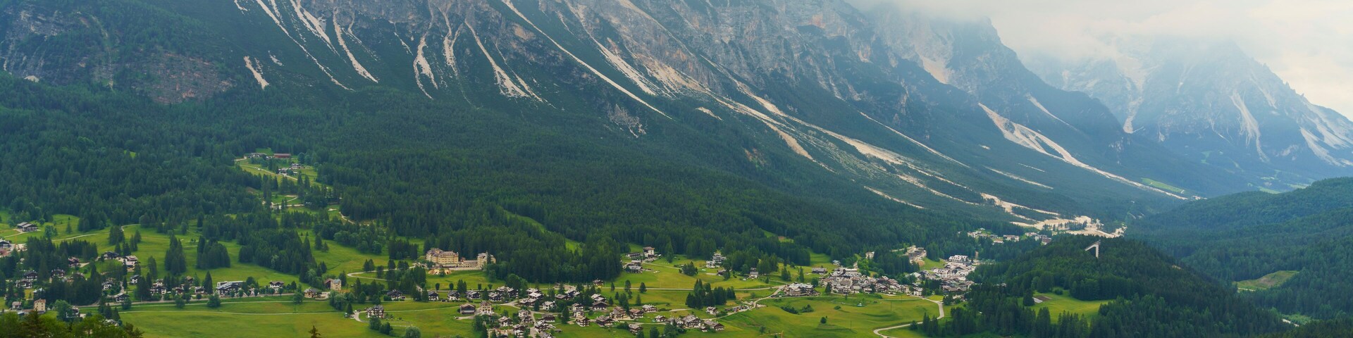 Paisagem montanhosa com uma aldeia aninhada num vale, rodeada por florestas densas e picos nevados.