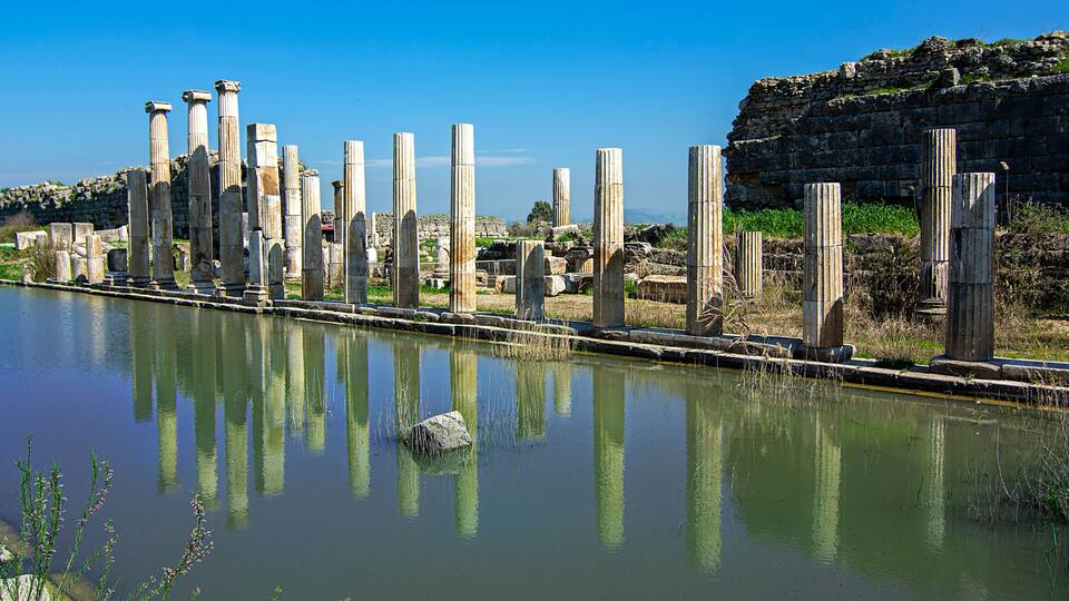 Ancient stone columns by a calm river, with a rocky cliff in the background.