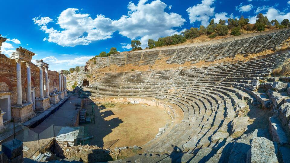 An ancient amphitheater with stone seating and columns, surrounded by trees and rocky terrain.