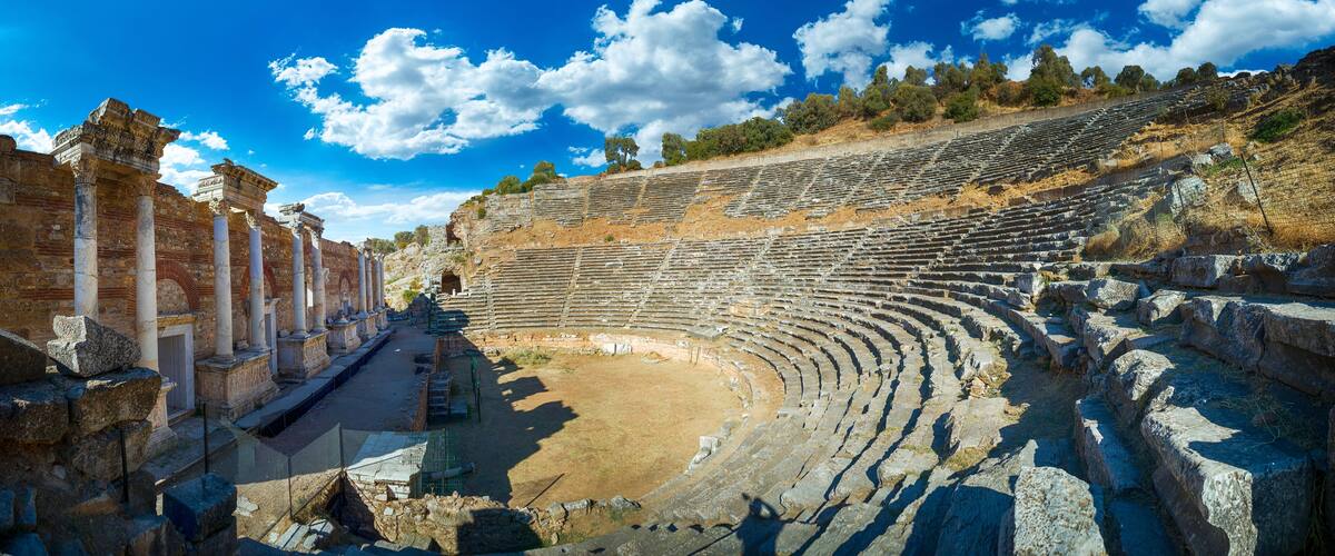An ancient amphitheater with stone seating and columns, surrounded by trees and rocky terrain.