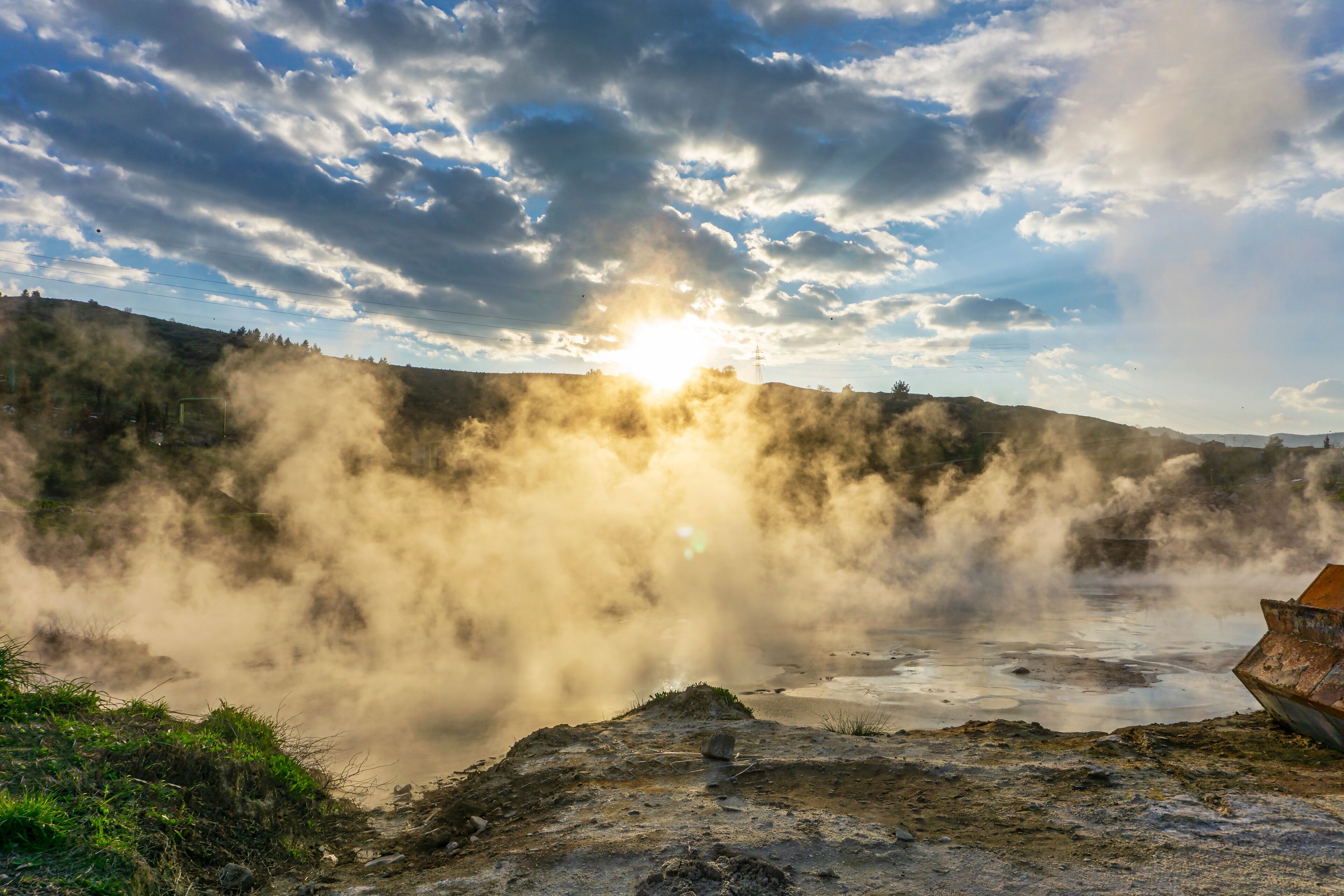 A geothermal area with steam rising from the ground, a boat on a rocky shore, and a cloudy sky.
