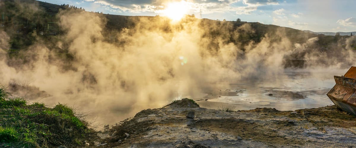 A geothermal area with steam rising from the ground, a boat on a rocky shore, and a cloudy sky.