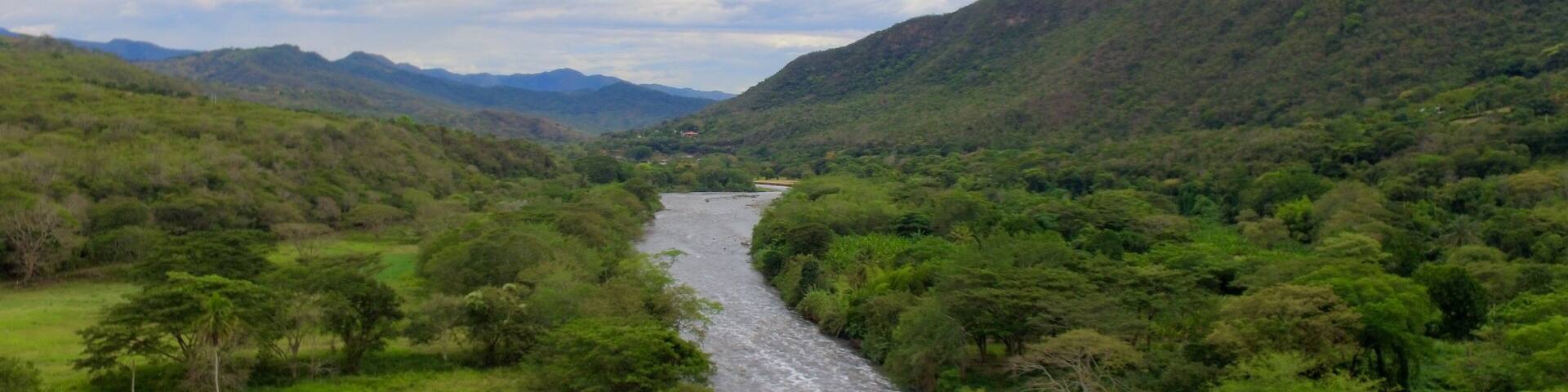 Wide aerial drone shot of the Río Bogotá cutting through a dense, jungle-like valley with steep, forested mountains on both sides near Apulo, Cundinamarca, Colombia.