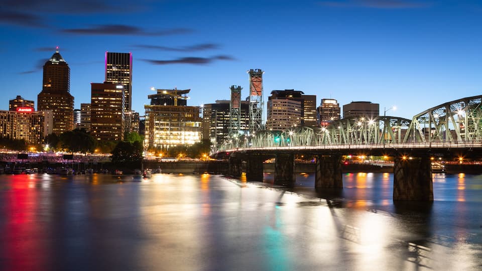 A city skyline at dusk with illuminated buildings and a bridge over a calm river.