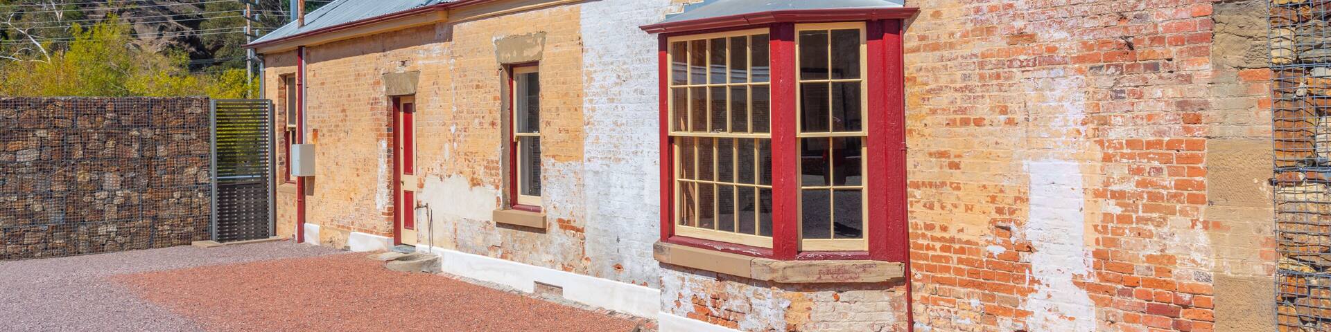 Un edificio histórico de ladrillo con detalles en rojo y un techo de metal, rodeado por un camino de grava y un muro de piedra.