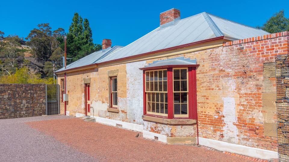 A historic brick building with red trim and a metal roof, surrounded by a gravel driveway and a stone wall.