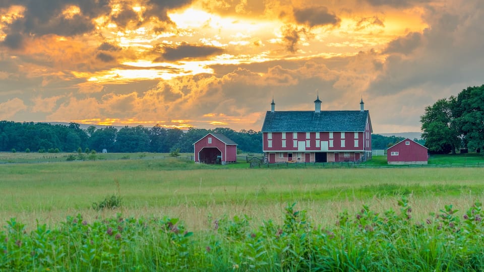 A red barn with a white fence in a grassy field under a sunset sky.