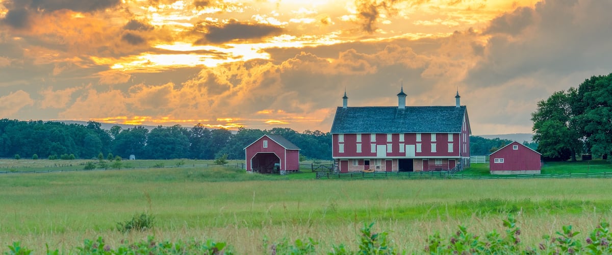 A red barn with a white fence in a grassy field under a sunset sky.