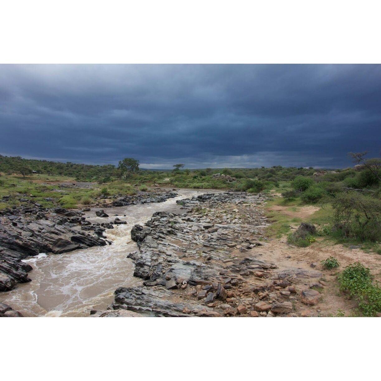 A rocky riverbed with a flowing stream, surrounded by green vegetation and a cloudy sky.