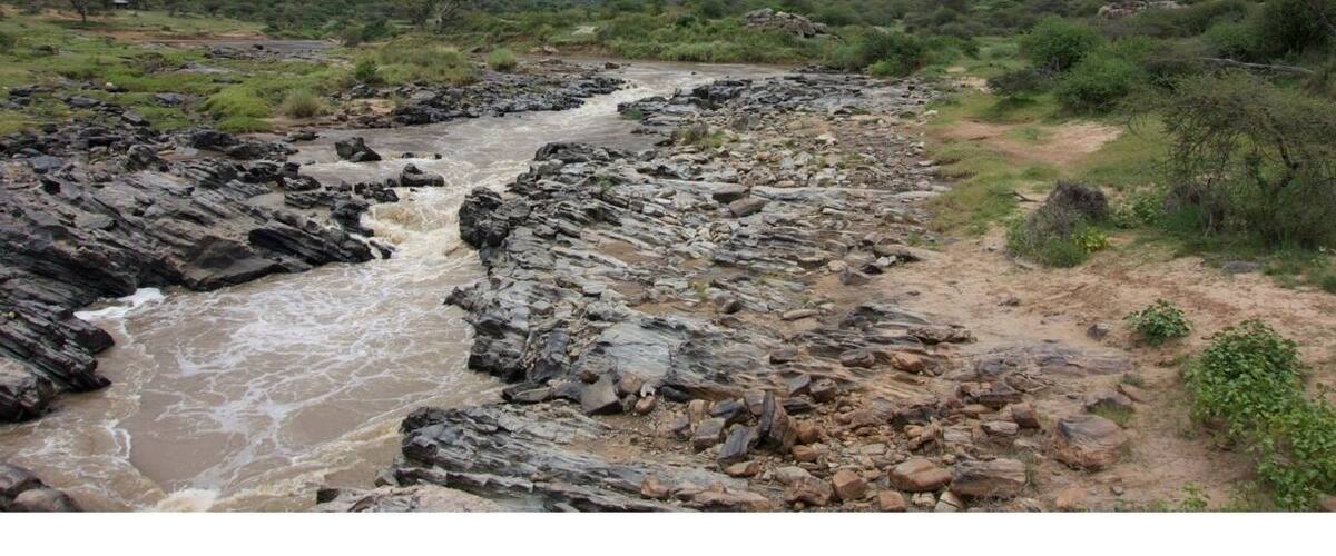 A rocky riverbed with a flowing stream, surrounded by green vegetation and a cloudy sky.