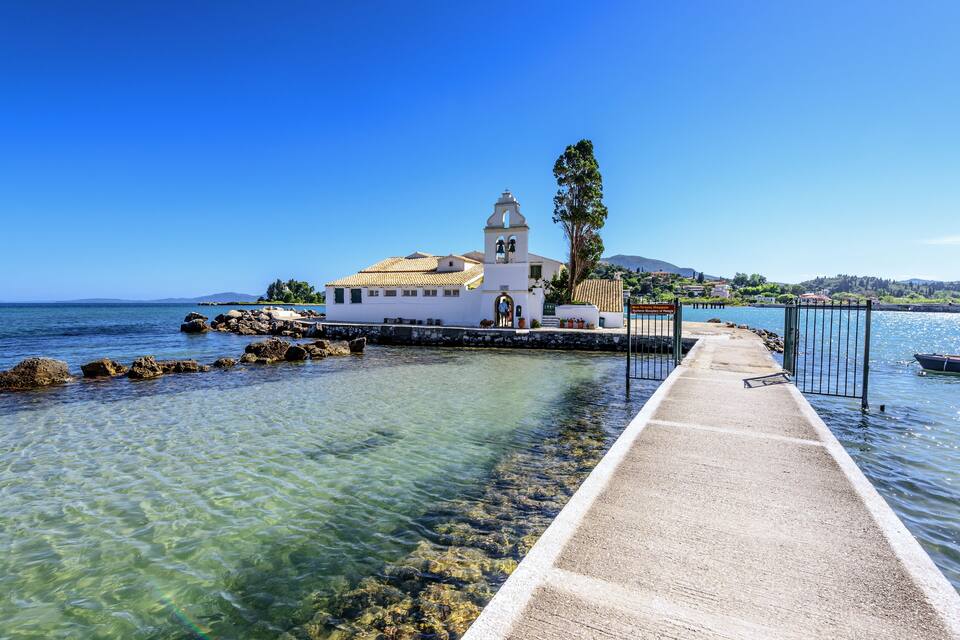 A coastal church with a bell tower, clear waters, and a pier leading to it.