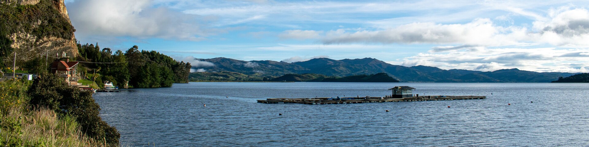A lake with a wooden pier, a small house on the shore, and mountains in the background.