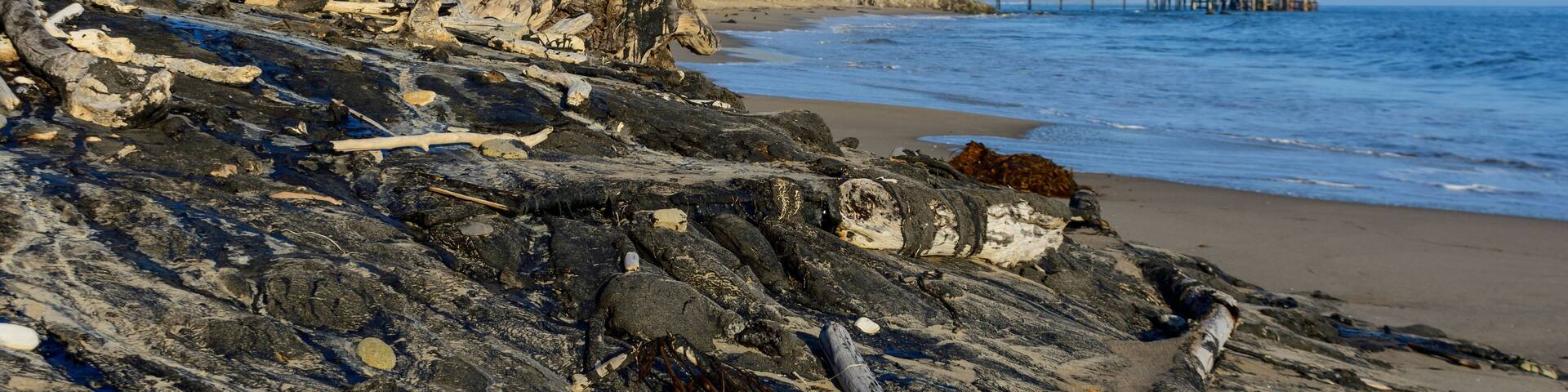 Coastline scenery at Tar Pits Park, Carpinteria, California, includes tar covered rocks and driftwood scattered along the shore. A pier stretches into the sea, set against a backdrop of coastal cliffs