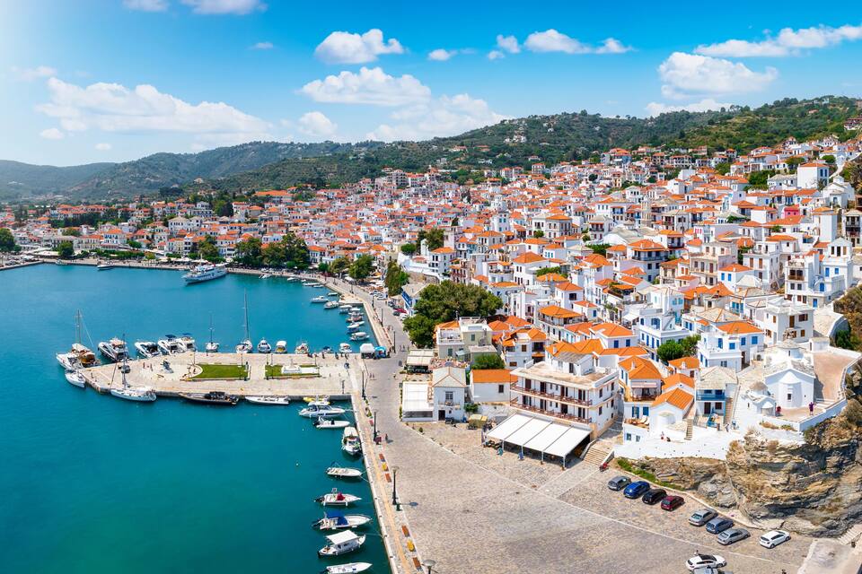 A coastal town with white buildings and red roofs, a marina, and a rocky coastline.