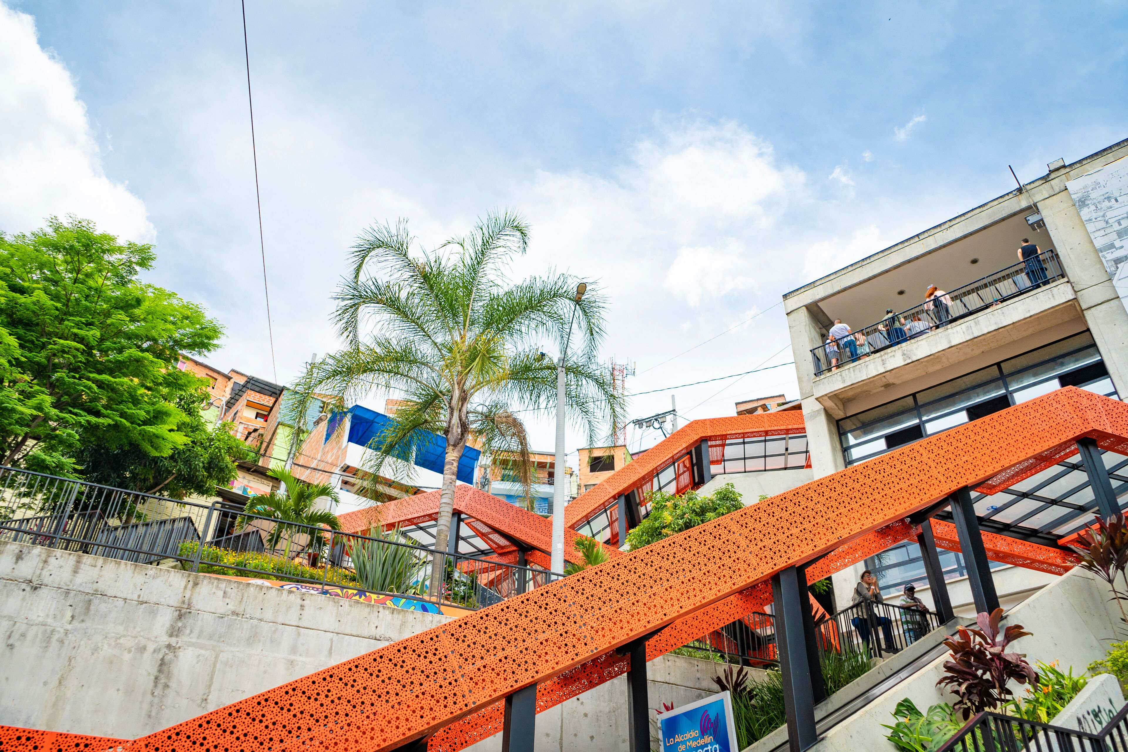A multi-level outdoor staircase with orange railings, surrounded by buildings and palm trees.