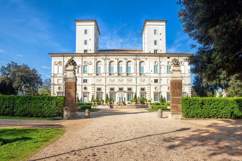 A large, historic building with two towers, surrounded by well-manicured gardens and a gravel pathway.