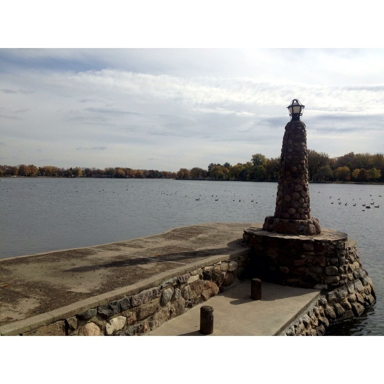 A small lighthouse made of stone rests upon the shores of Black Hawk Lake, where flocks of wood ducks and Canadian geese are swimming around.