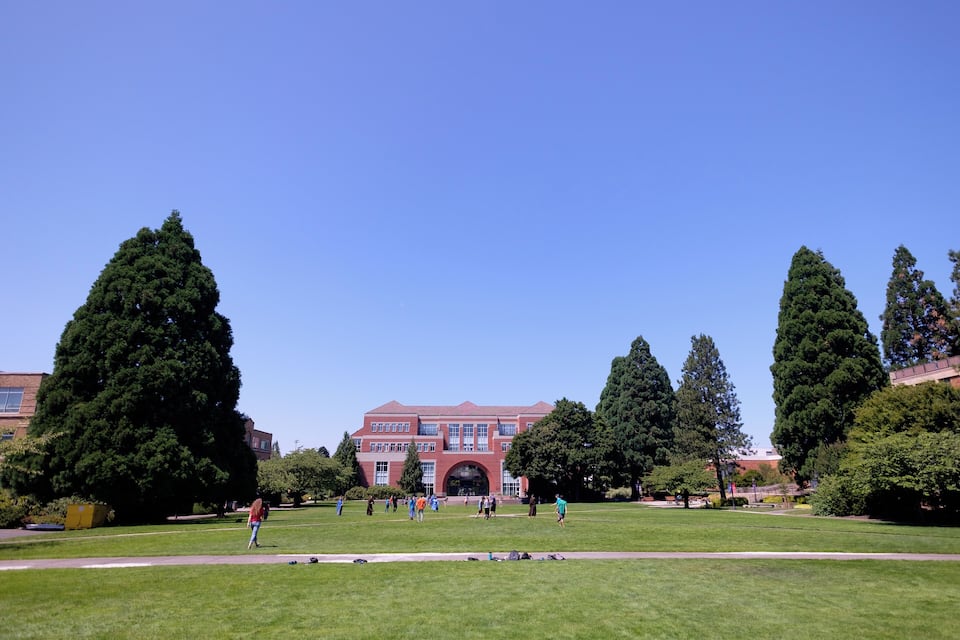 A large red building with a central archway, surrounded by a grassy field and tall trees.