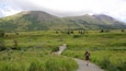 A hiker on a gravel path with a wooden bridge, surrounded by green hills and a mountain in the background.