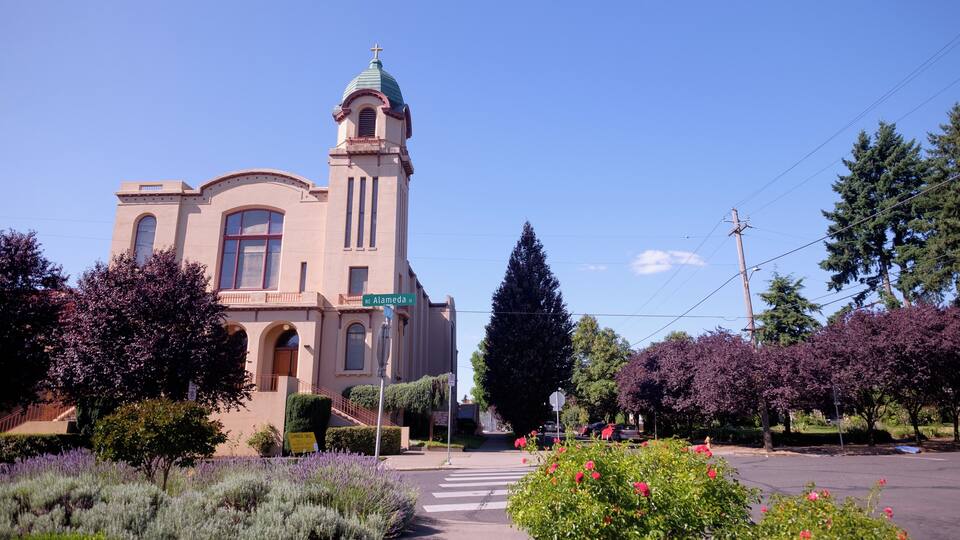 A church with a tall tower and arched windows, surrounded by flowering bushes and trees.